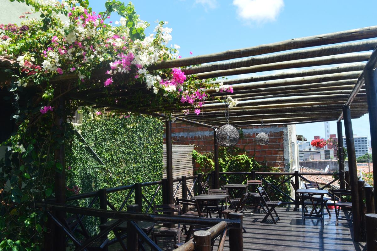 Terraço com deck de madeira, mesas e pergolado coberto por buganvília